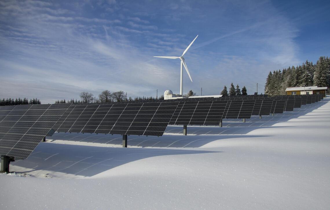 Solar panels in the snow with a windmill under a sunny sky.